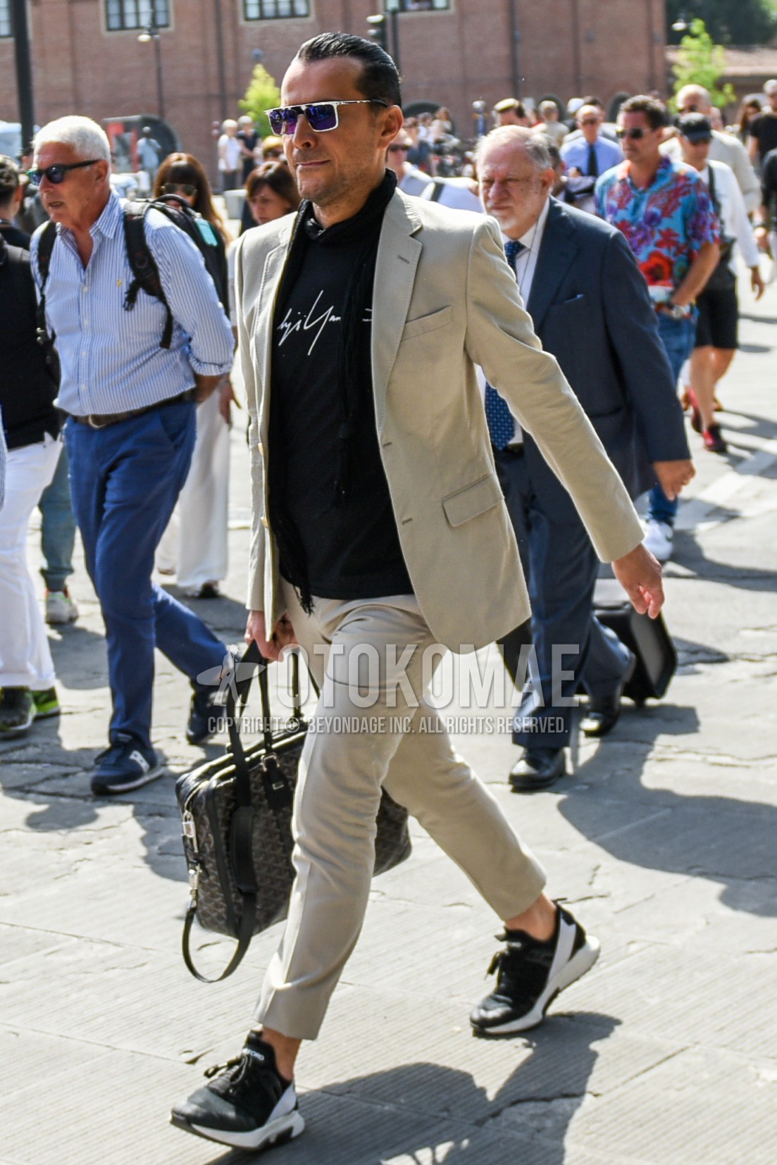 Men's spring autumn outfit with white plain sunglasses, black plain scarf, black deca logo t-shirt, black low-cut sneakers, graphic briefcase/handbag, beige plain suit.