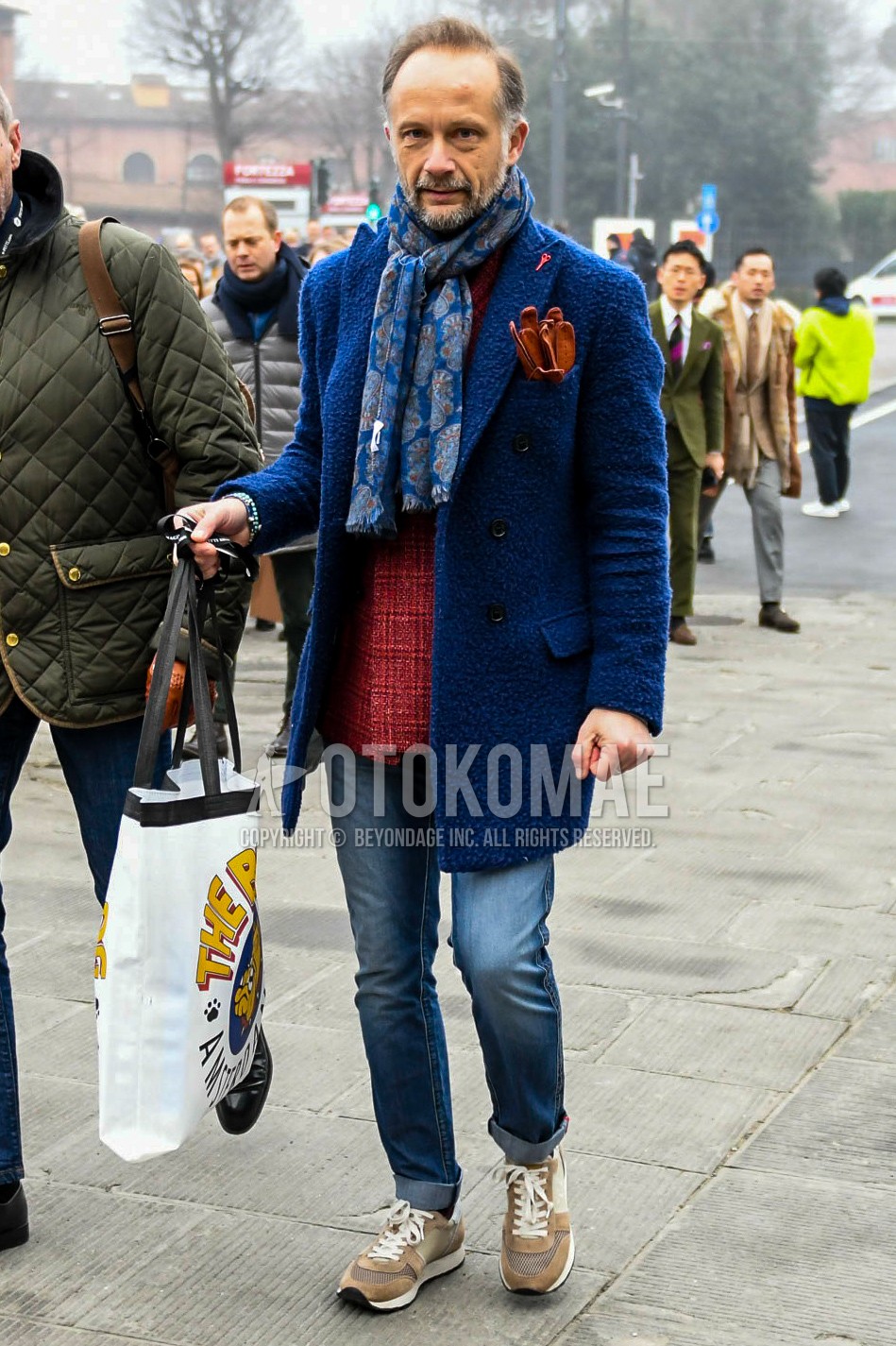 Men's winter outfit with blue scarf scarf, blue plain chester coat, red check tailored jacket, blue plain denim/jeans, brown low-cut sneakers.