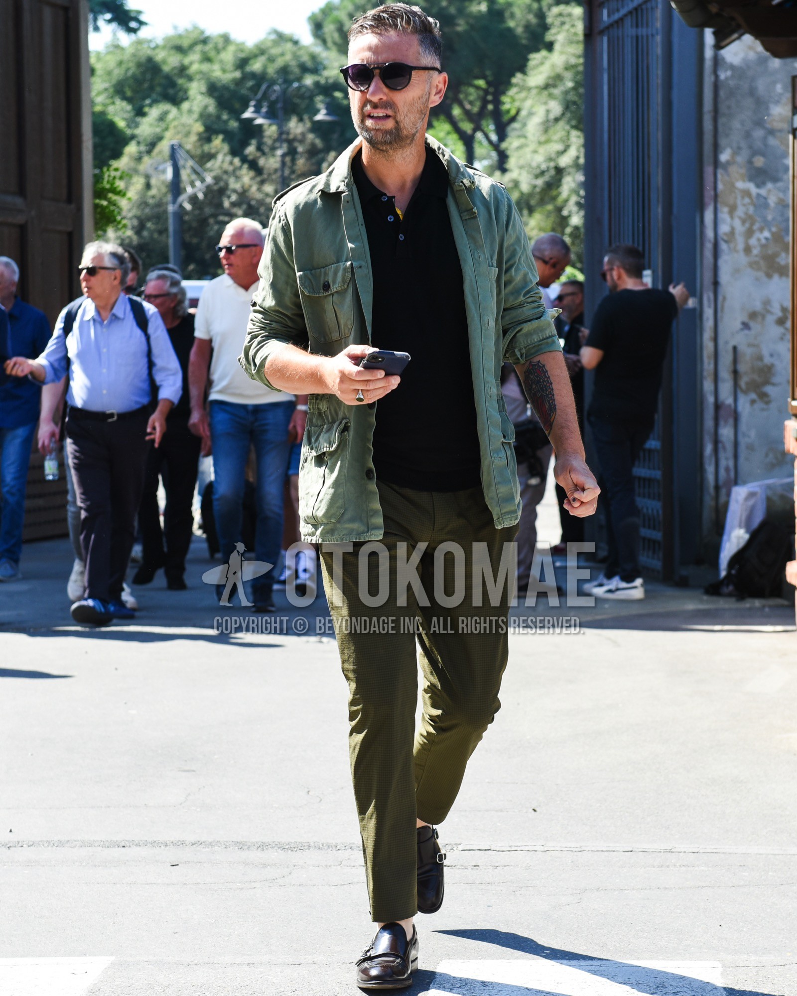 Men's spring summer outfit with black plain sunglasses, olive green plain field jacket/hunting jacket, black plain polo shirt, olive green check ankle pants, brown monk shoes leather shoes.