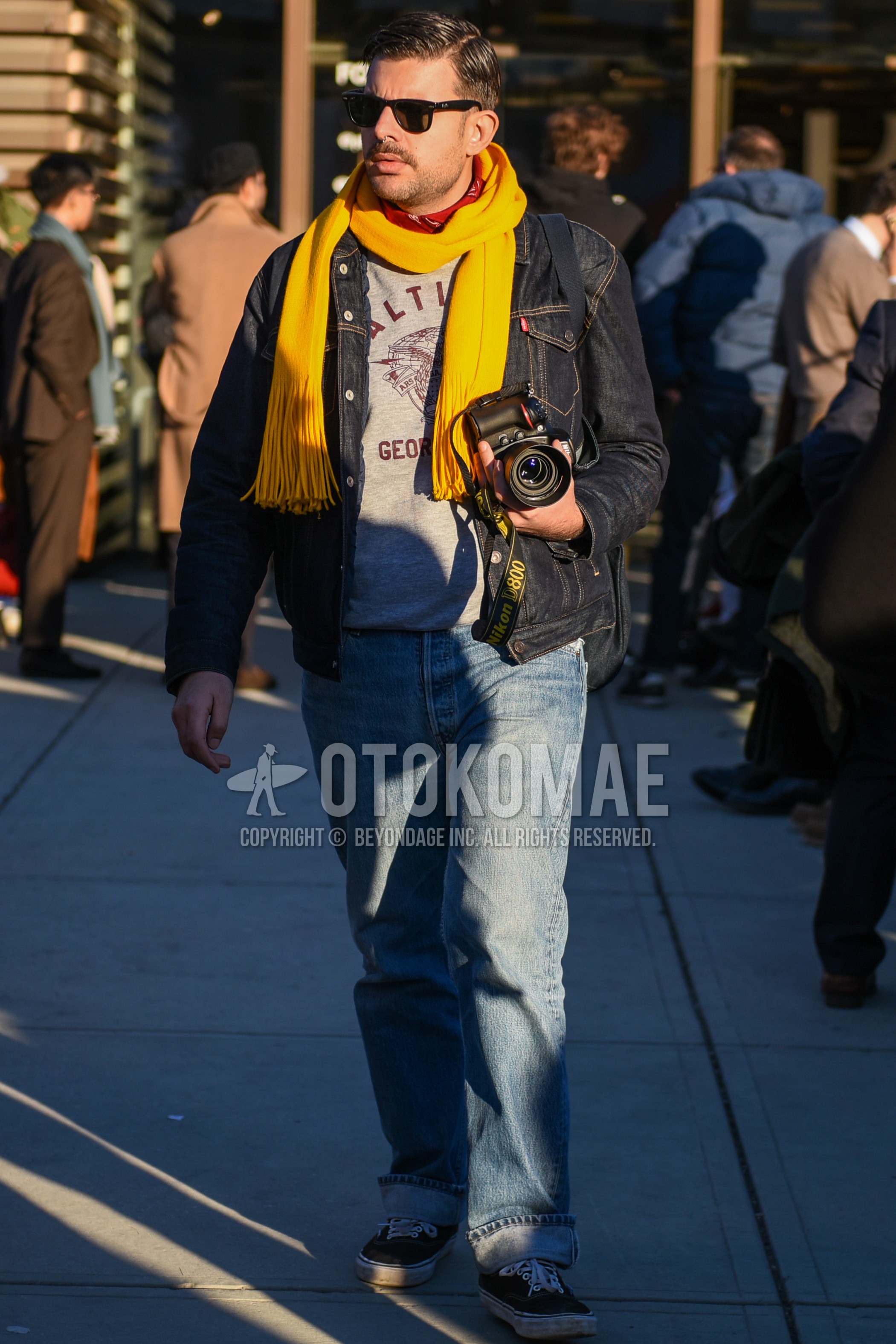 Men's autumn winter outfit with black plain sunglasses, yellow plain scarf, navy plain denim jacket, gray graphic sweatshirt, blue plain denim/jeans, black low-cut sneakers.