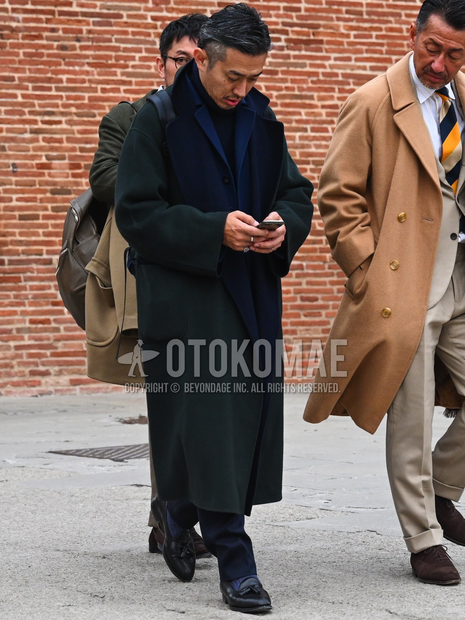 Men's autumn winter outfit with green plain ulster coat, navy plain tailored jacket, black plain turtleneck knit, navy plain slacks, navy plain socks, black tassel loafers leather shoes, black plain backpack.