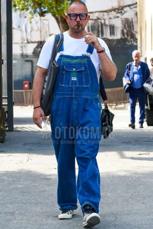 黒無地のサングラス、オーバーオールのブルー無地のジャンプスーツ、白ワンポイントのTシャツ、白ローカットのスニーカー、ダークグレーグラフィックのトートバッグを合わせた夏のメンズコーデ・着こなし。