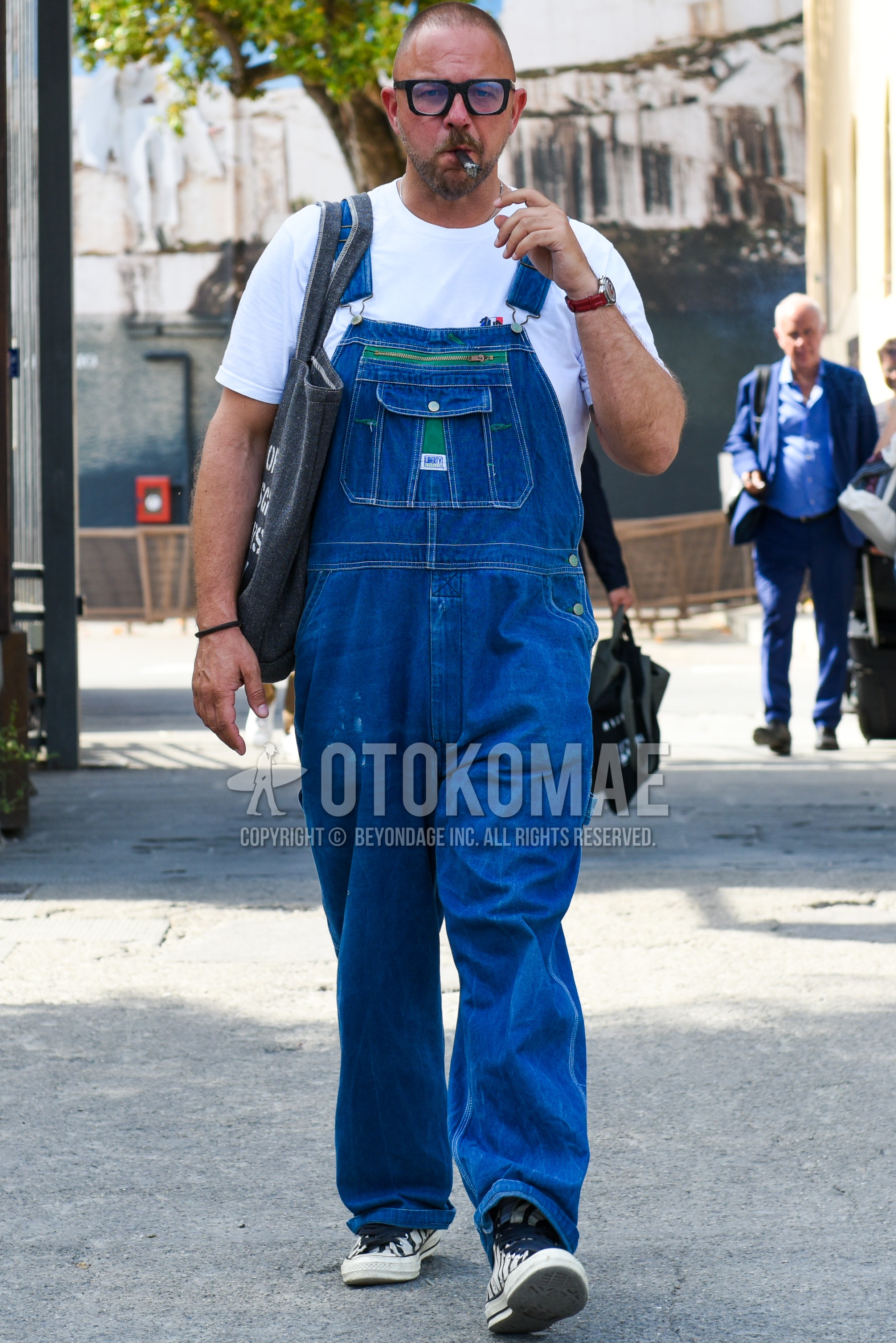 Men's summer outfit with black plain sunglasses, blue plain jumpsuit, white one point t-shirt, white low-cut sneakers, dark gray graphic tote bag.