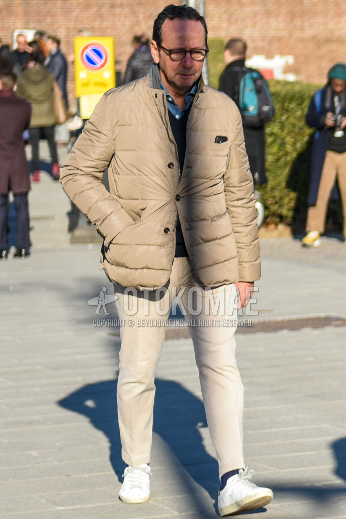 Men's autumn winter outfit with brown tortoiseshell glasses, beige plain tailored jacket, beige plain down jacket, black plain sweater, blue plain denim shirt/chambray shirt, beige plain chinos, beige plain ankle pants, gray plain socks, white low-cut sneakers.