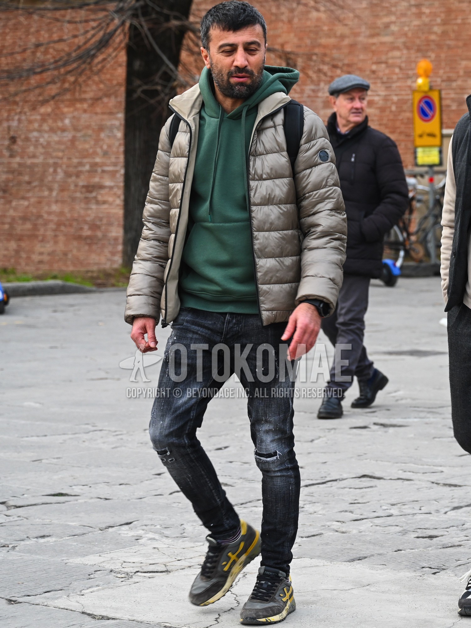 Men's autumn winter outfit with beige plain down jacket, green plain hoodie, black plain damaged jeans, black plain denim/jeans, gray low-cut sneakers.