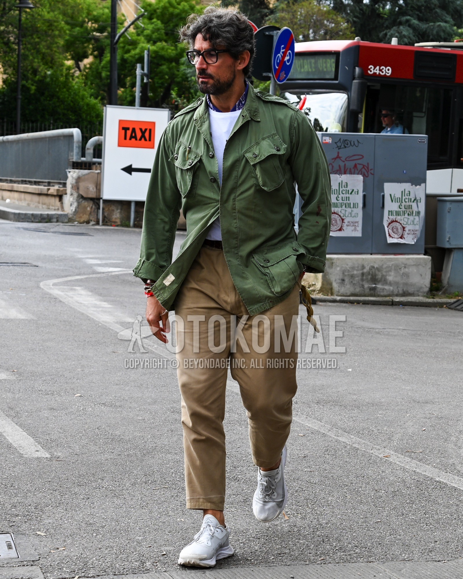 Men's spring summer autumn outfit with clear plain sunglasses, black scarf bandana/neckerchief, olive green plain military jacket, white plain t-shirt, beige plain chinos, white low-cut sneakers.