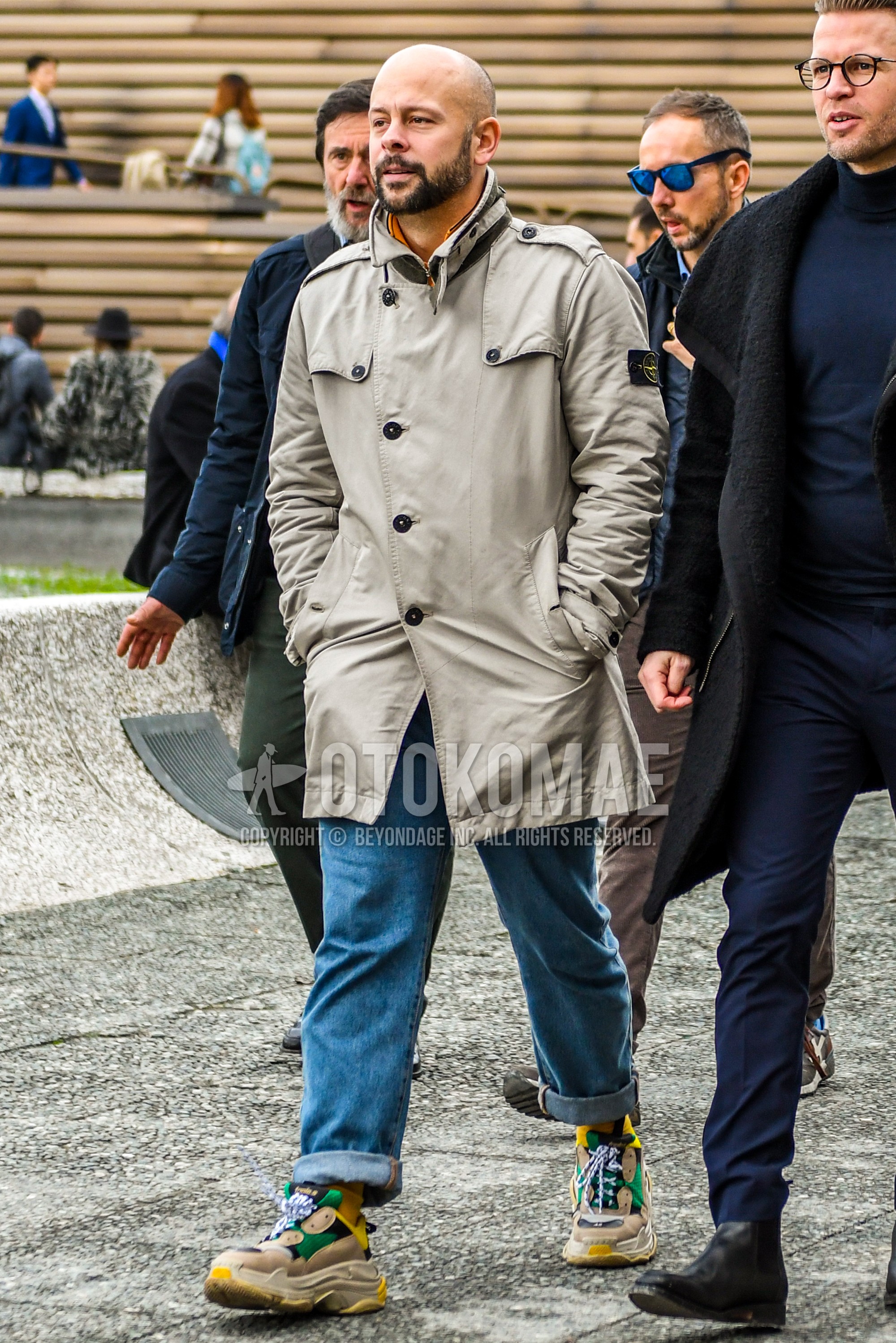 Men's autumn winter outfit with gray plain trench coat, blue plain denim/jeans, multi-color green low-cut sneakers.