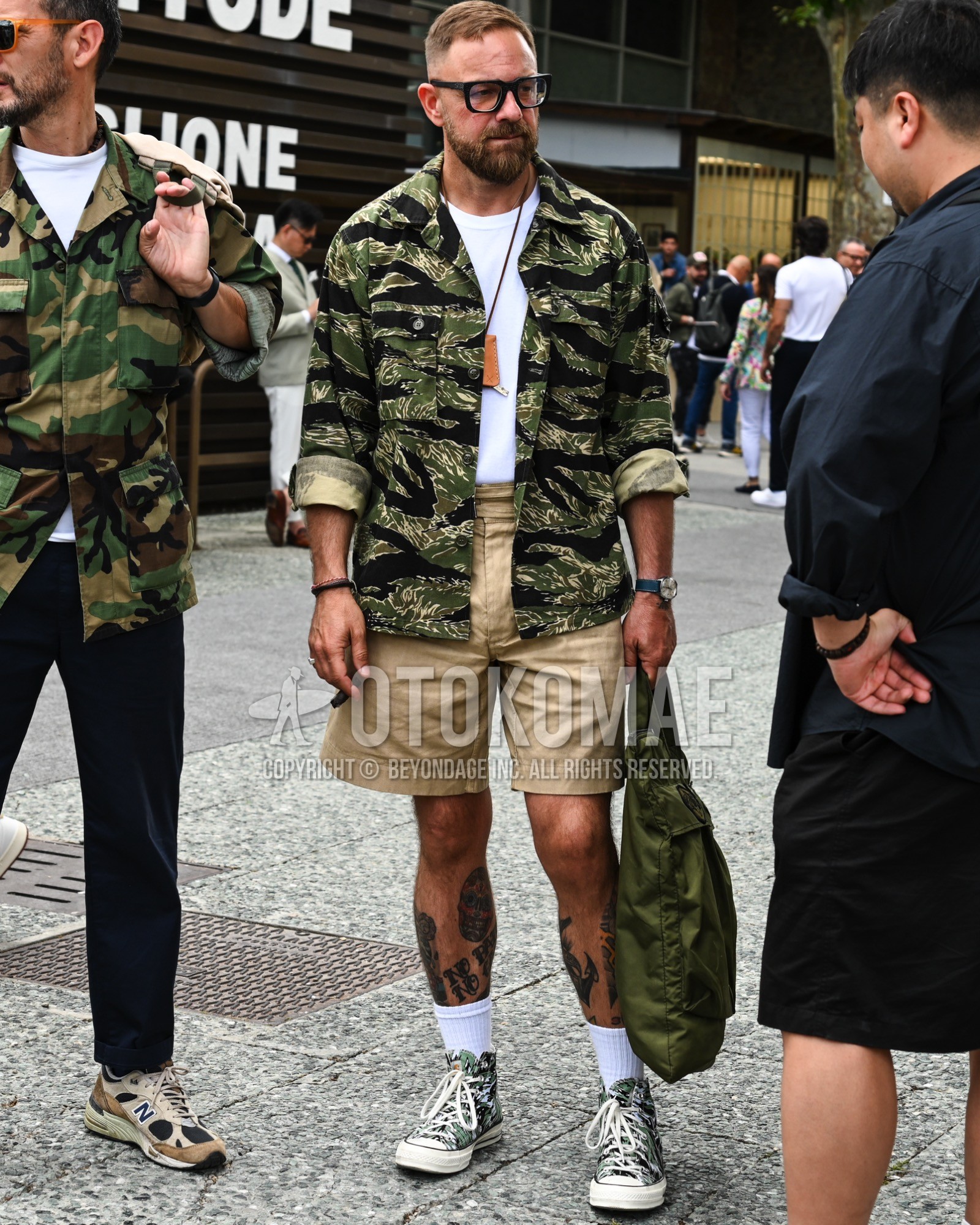 Men's spring summer outfit with clear plain sunglasses, olive green camouflage military jacket, white plain t-shirt, beige plain short pants, white plain socks, green low-cut sneakers, green plain tote bag.