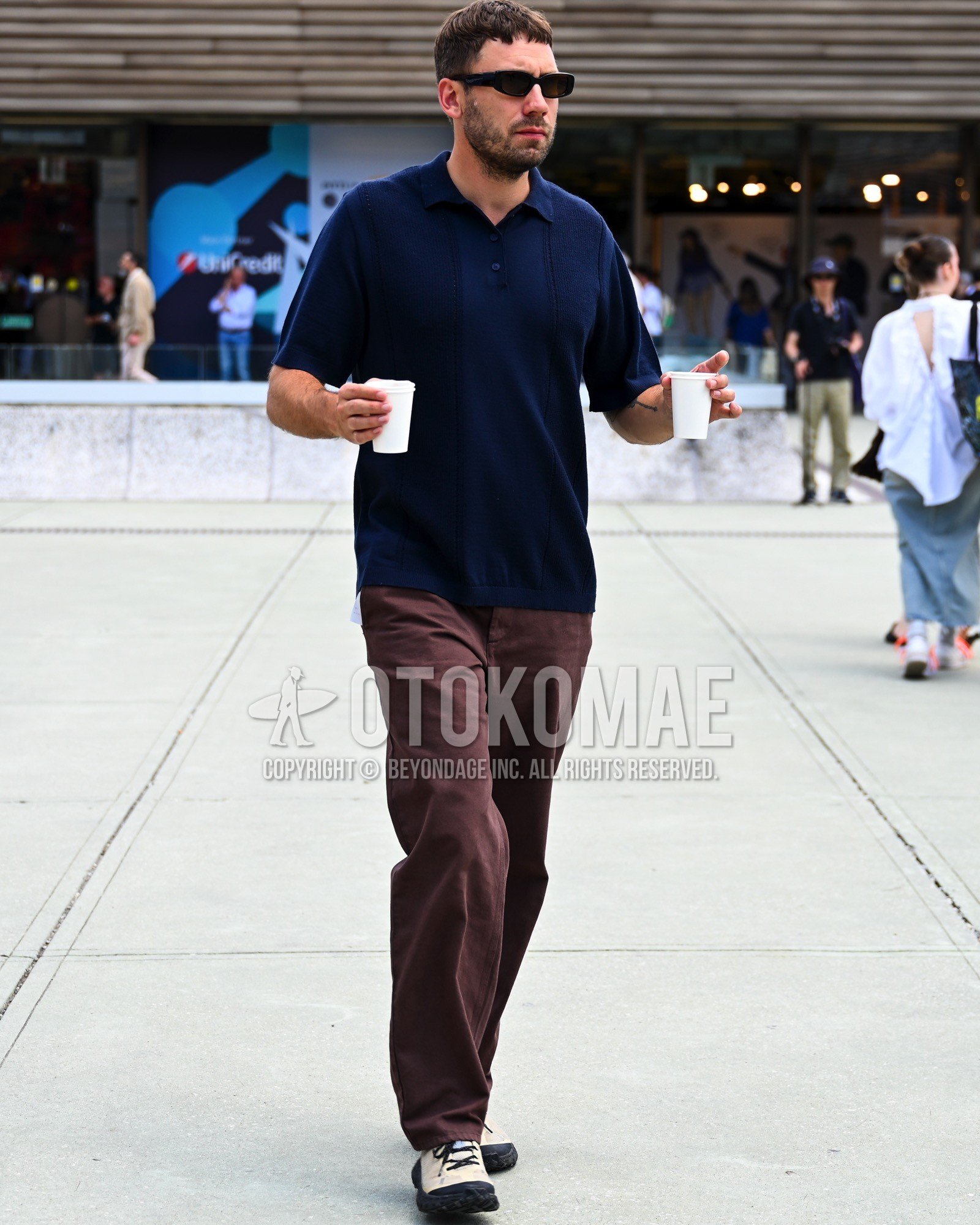 Men's spring summer outfit with black plain sunglasses, navy plain polo shirt, brown plain chinos, white low-cut sneakers.