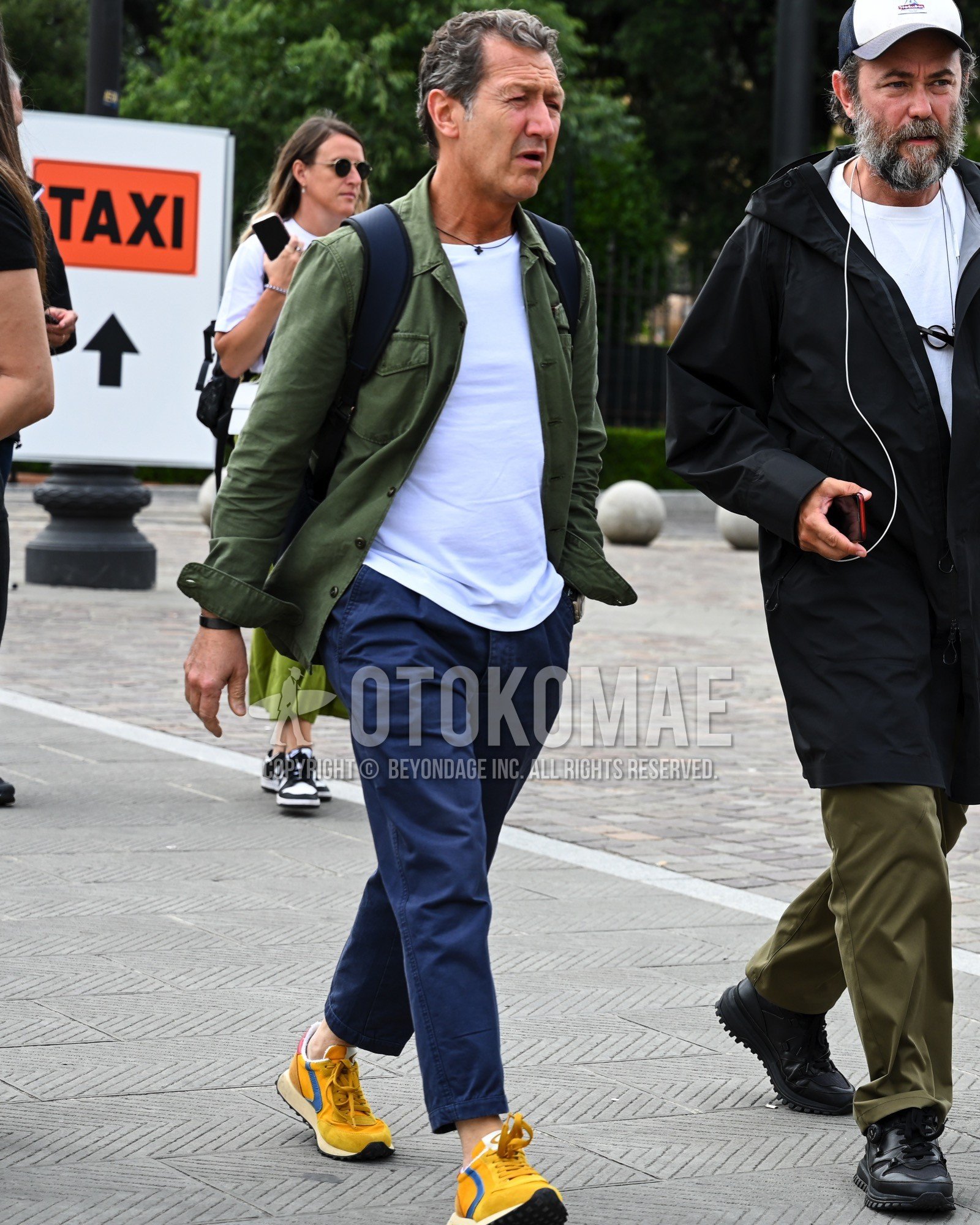 Men's spring summer autumn outfit with olive green plain military jacket, white plain t-shirt, navy plain chinos, yellow low-cut sneakers, black plain backpack.