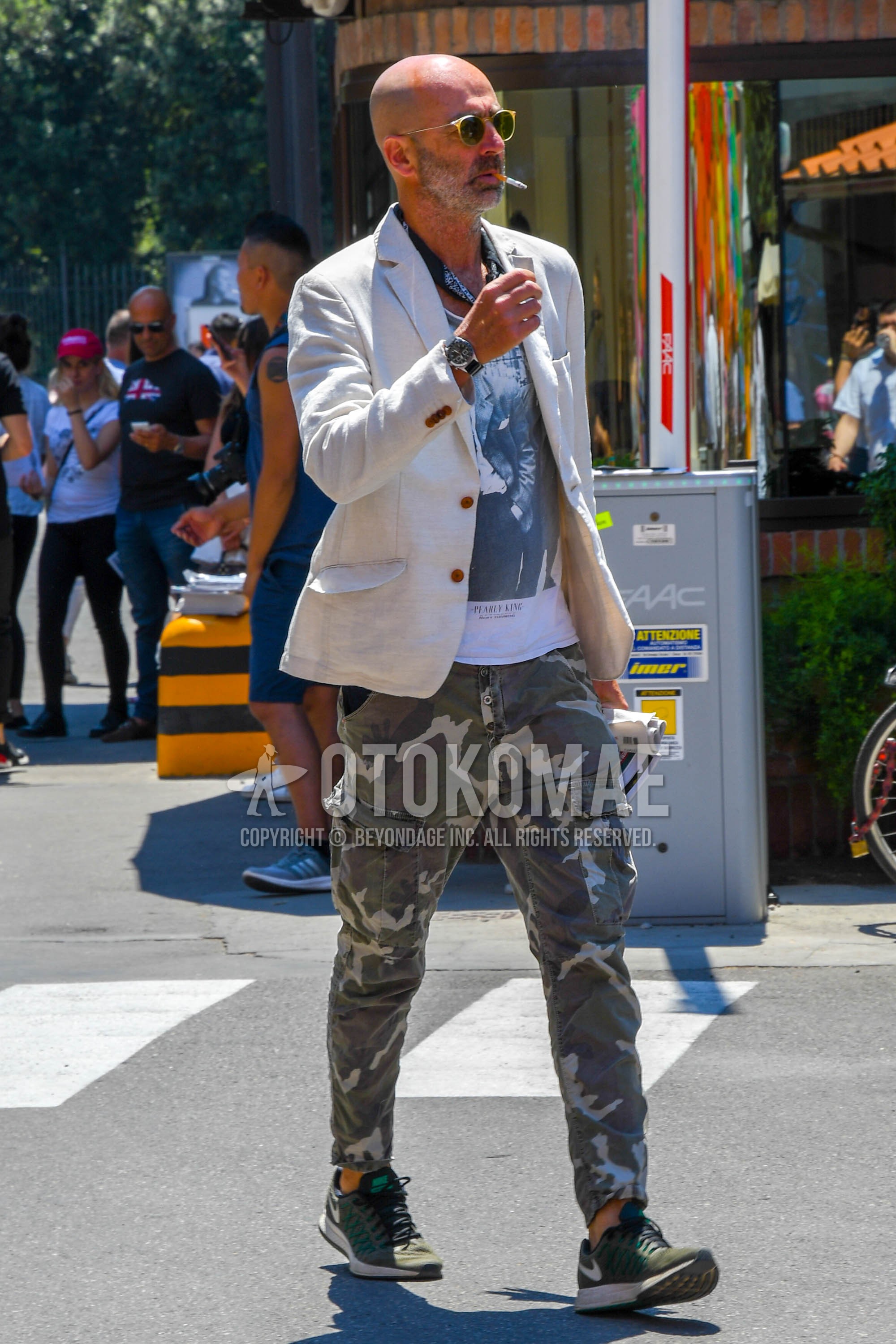 Men's spring summer autumn outfit with plain sunglasses, beige plain tailored jacket, white graphic t-shirt, white olive green camouflage cargo pants, olive green low-cut sneakers.
