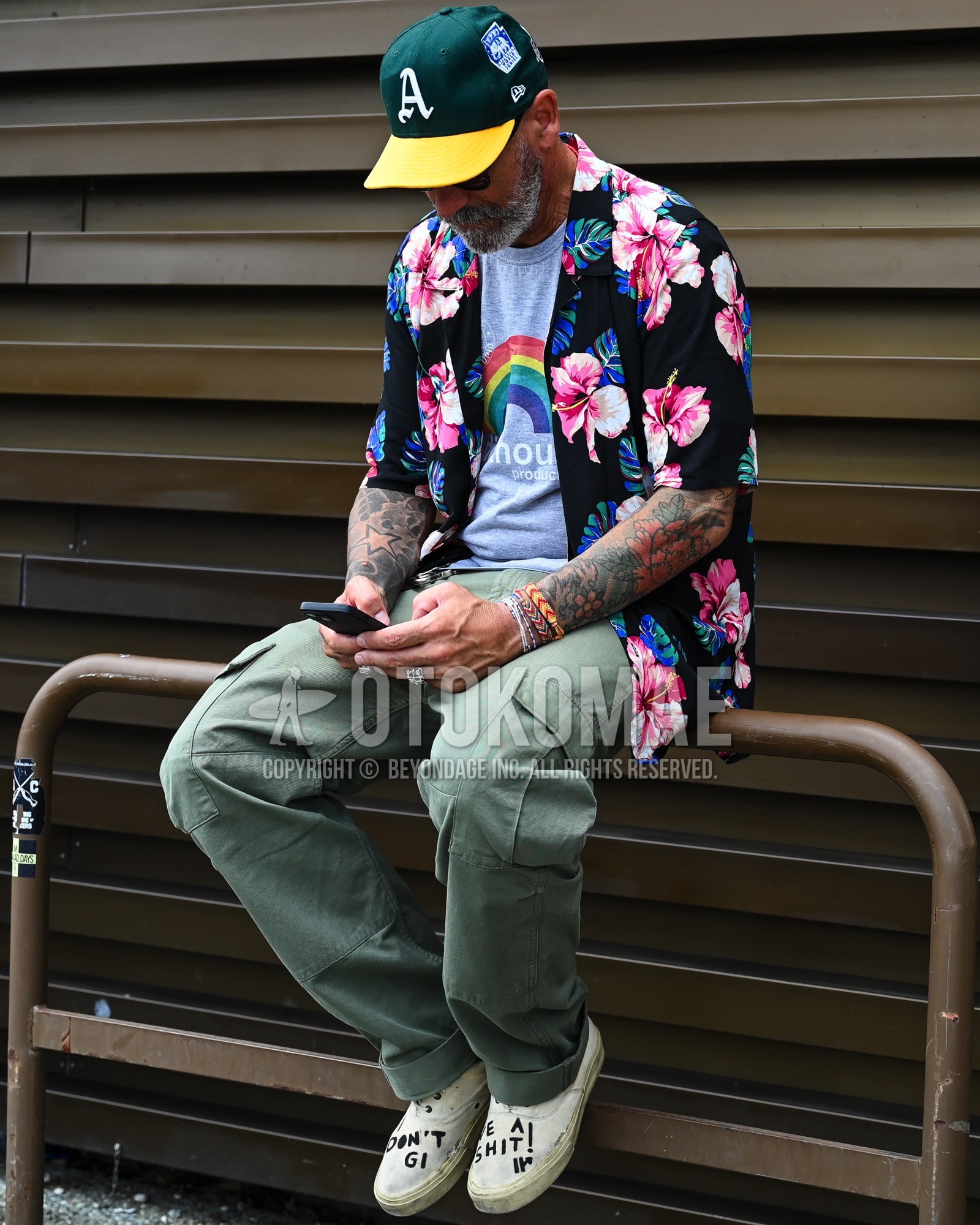 Men's spring summer outfit with green one point baseball cap, black whole pattern shirt, gray tops/innerwear t-shirt, olive green plain cargo pants, white slip-on sneakers.