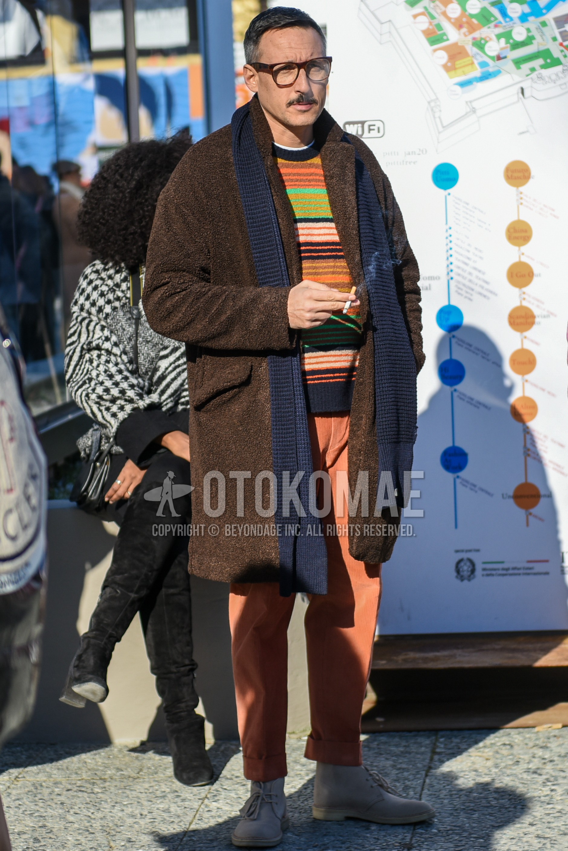 Men's autumn winter outfit with black plain glasses, gray plain scarf, brown plain chester coat, multi-color horizontal stripes sweater, orange plain winter pants (corduroy,velour), beige chukka boots.