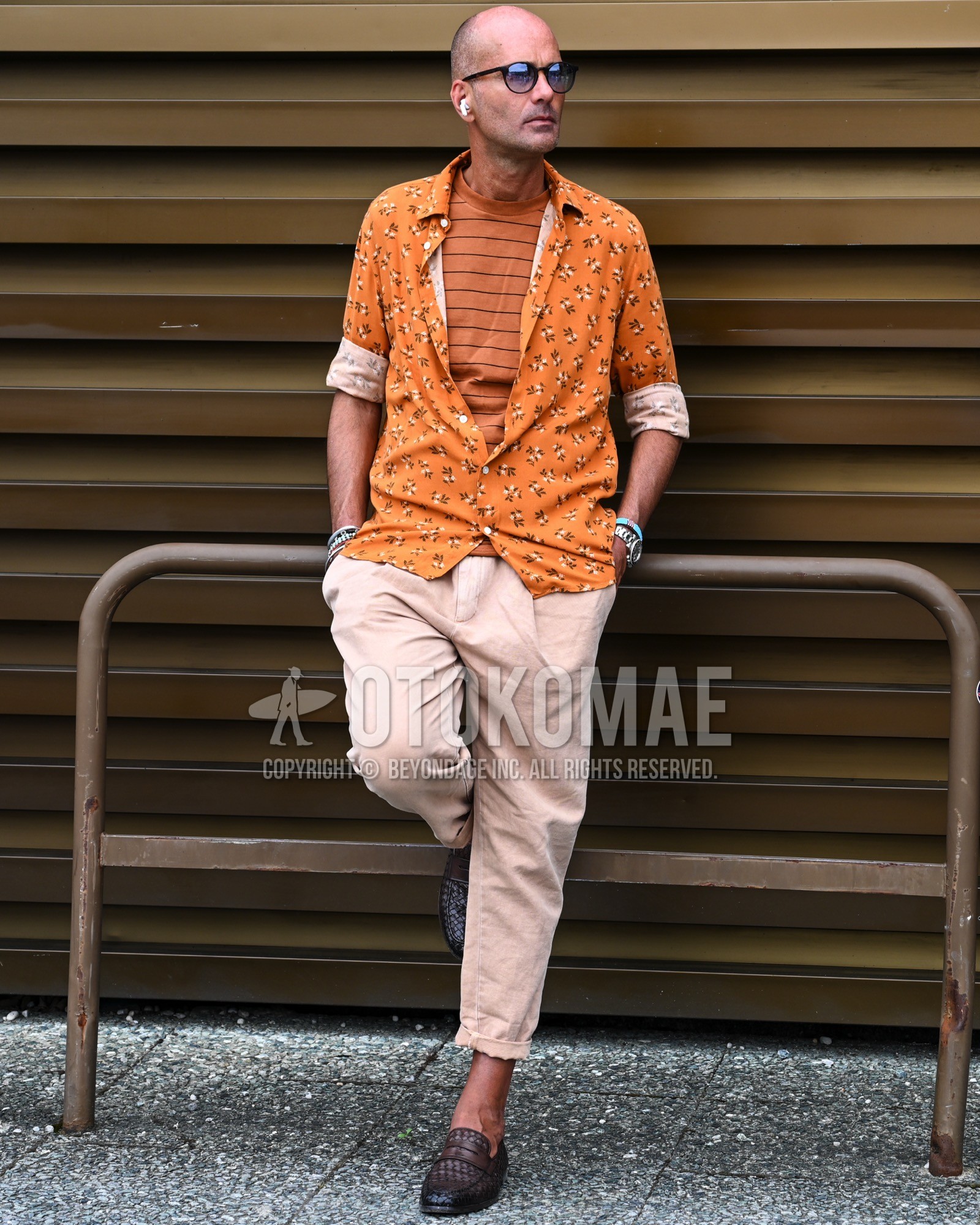 Men's spring summer autumn outfit with blue plain sunglasses, orange whole pattern shirt, orange horizontal stripes t-shirt, beige plain chinos, brown  loafers leather shoes.