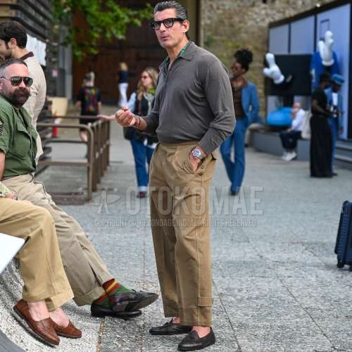 ウェリントンの黒無地のメガネ、黒ストールのスカーフ、グレー無地のポロシャツ、ベージュボトムスのスラックス、ベージュボトムスのプリーツパンツ、ベージュボトムスのコットンパンツ、ブラウンローファーの革靴を合わせた夏春のメンズコーデ・着こなし。