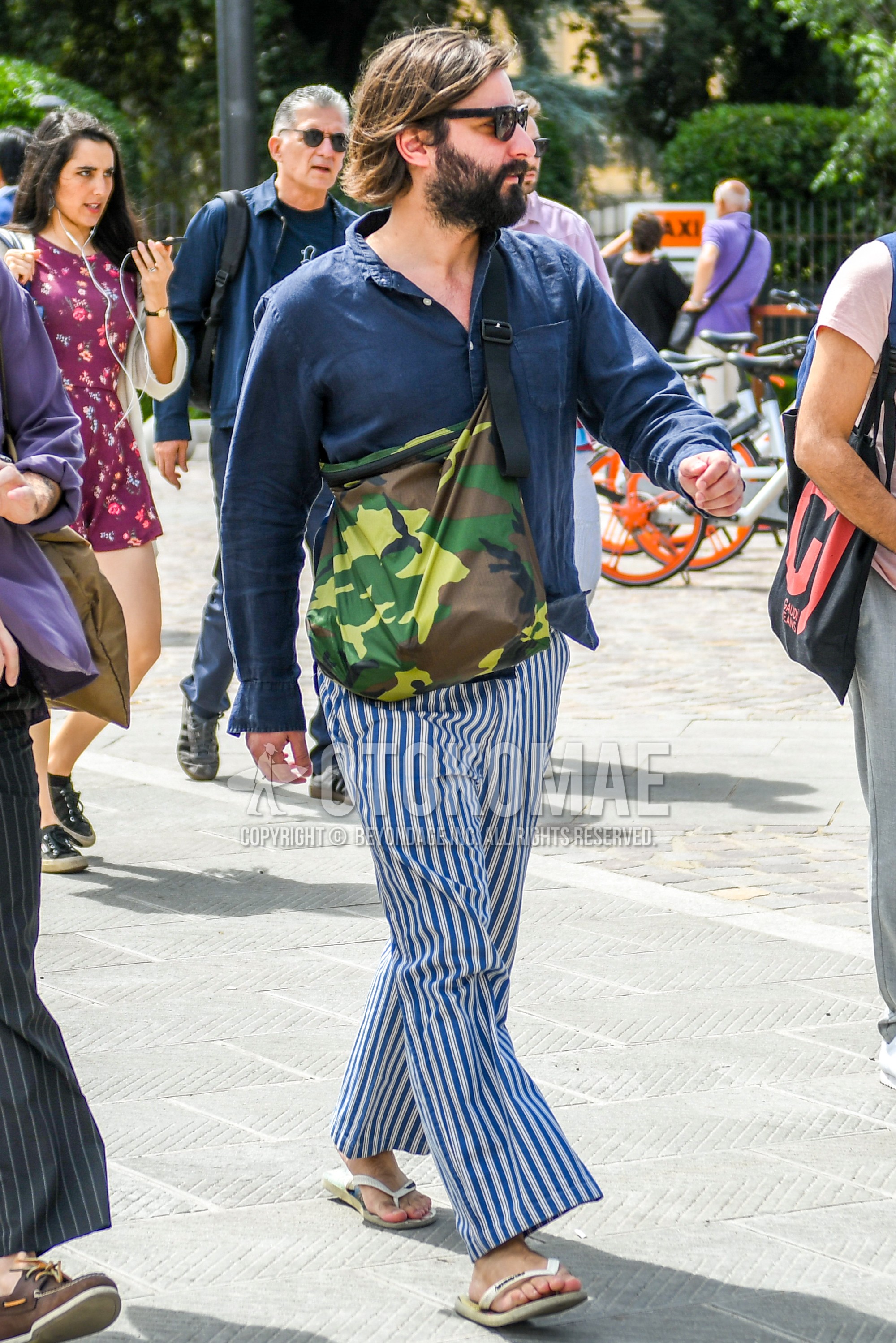 Men's spring summer autumn outfit with plain sunglasses, navy plain shirt, blue white stripes wide pants, white flip flops sandals, multi-color green camouflage shoulder bag.