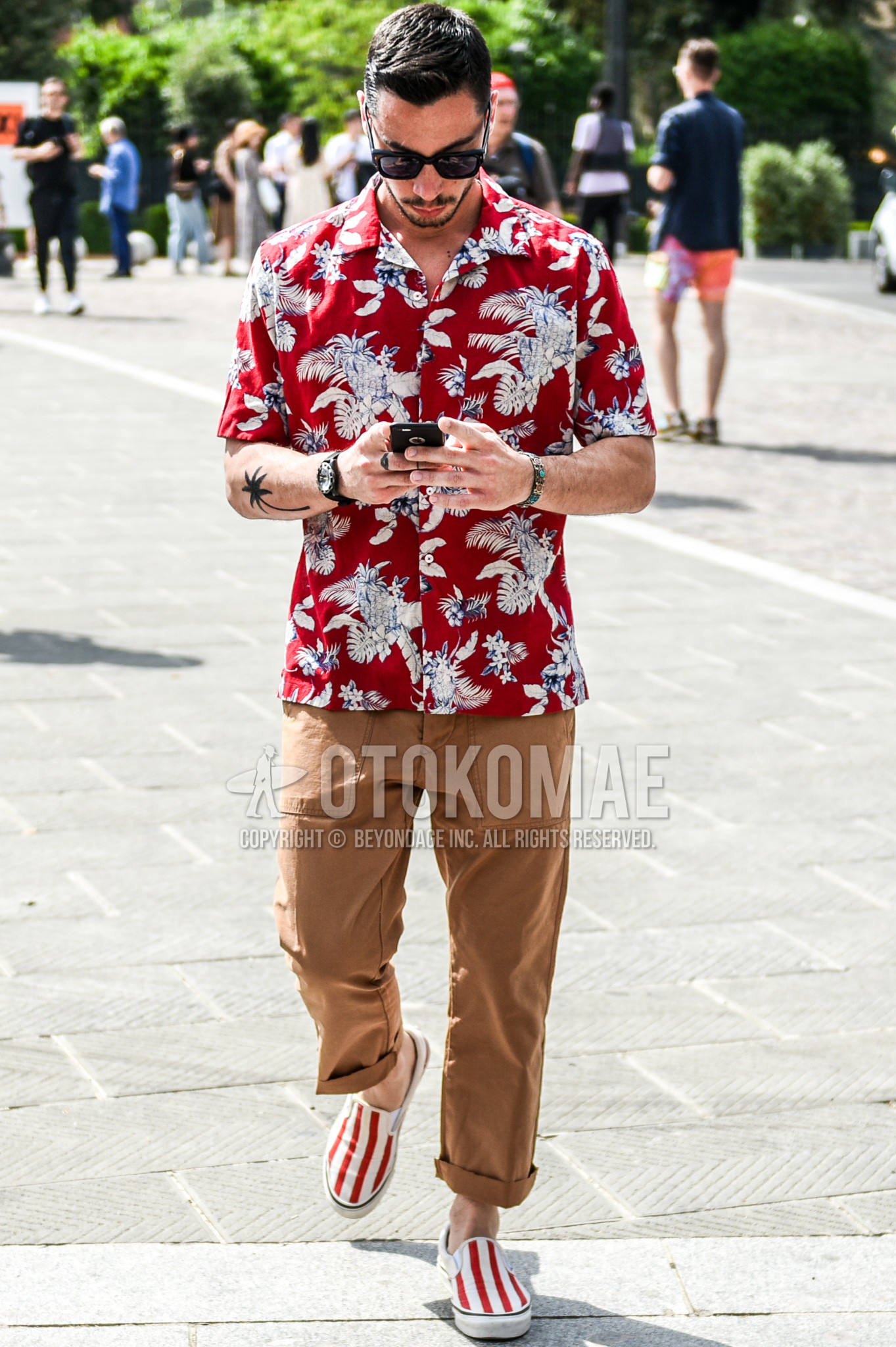 Men's summer outfit with black plain sunglasses, red botanical shirt, beige brown plain cargo pants, white red slip-on sneakers.