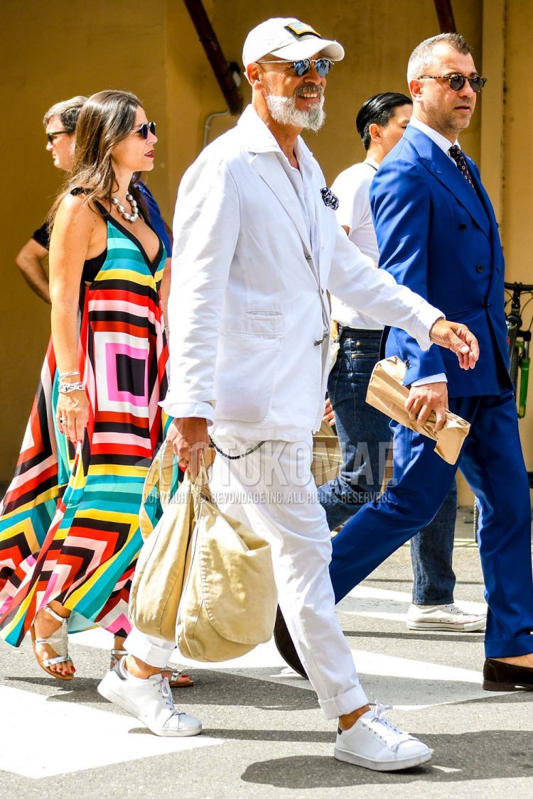 A summer-spring-fall men's outfit with a solid color baseball cap, solid color sunglasses, solid color white tailored jacket, solid color white shirt, solid color white cotton pants, white low-cut sneakers, and a solid color beige tote bag.
