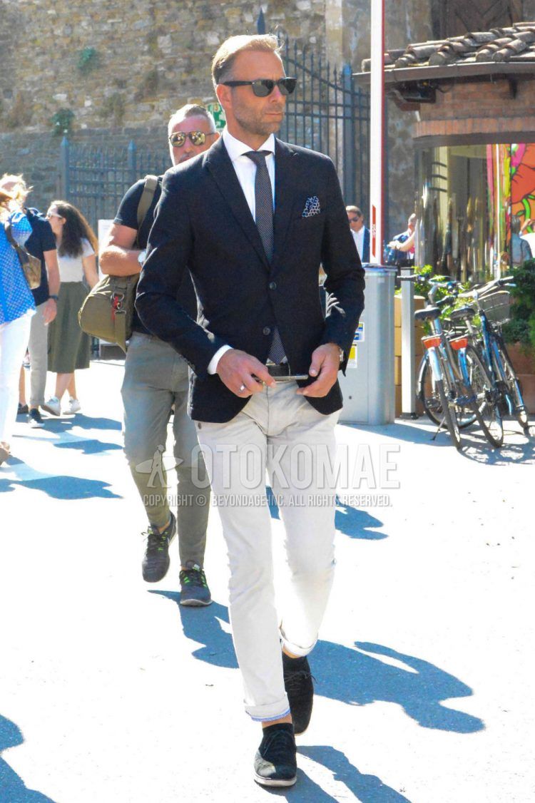 A men's spring/summer/fall outfit with solid color sunglasses, solid color black tailored jacket, solid color white shirt, solid color black leather belt, solid color beige chinos, black brogue shoes leather shoes, suede shoes leather shoes, and solid color gray tie.
