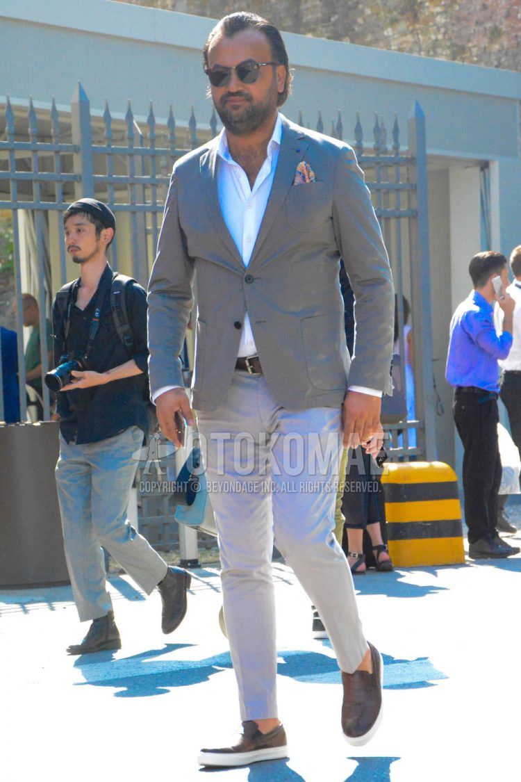Spring, summer and fall men's coordinate outfit with plain brown sunglasses, plain gray tailored jacket, plain white shirt, plain brown leather belt, plain beige chinos and brown low-cut sneakers.