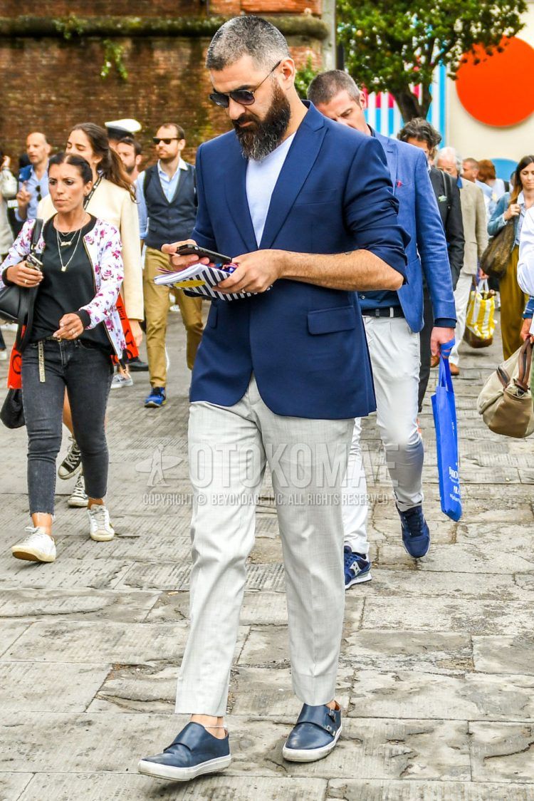 Spring, summer and fall men's coordinate outfit with plain gold sunglasses, plain blue tailored jacket, plain white t-shirt, plain gray slacks and blue slip-on sneakers.