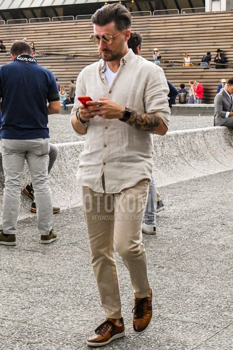 A spring/summer men's coordinate outfit with solid color sunglasses, solid color beige shirt, solid color white t-shirt, solid color beige chinos, and brown low-cut sneakers.