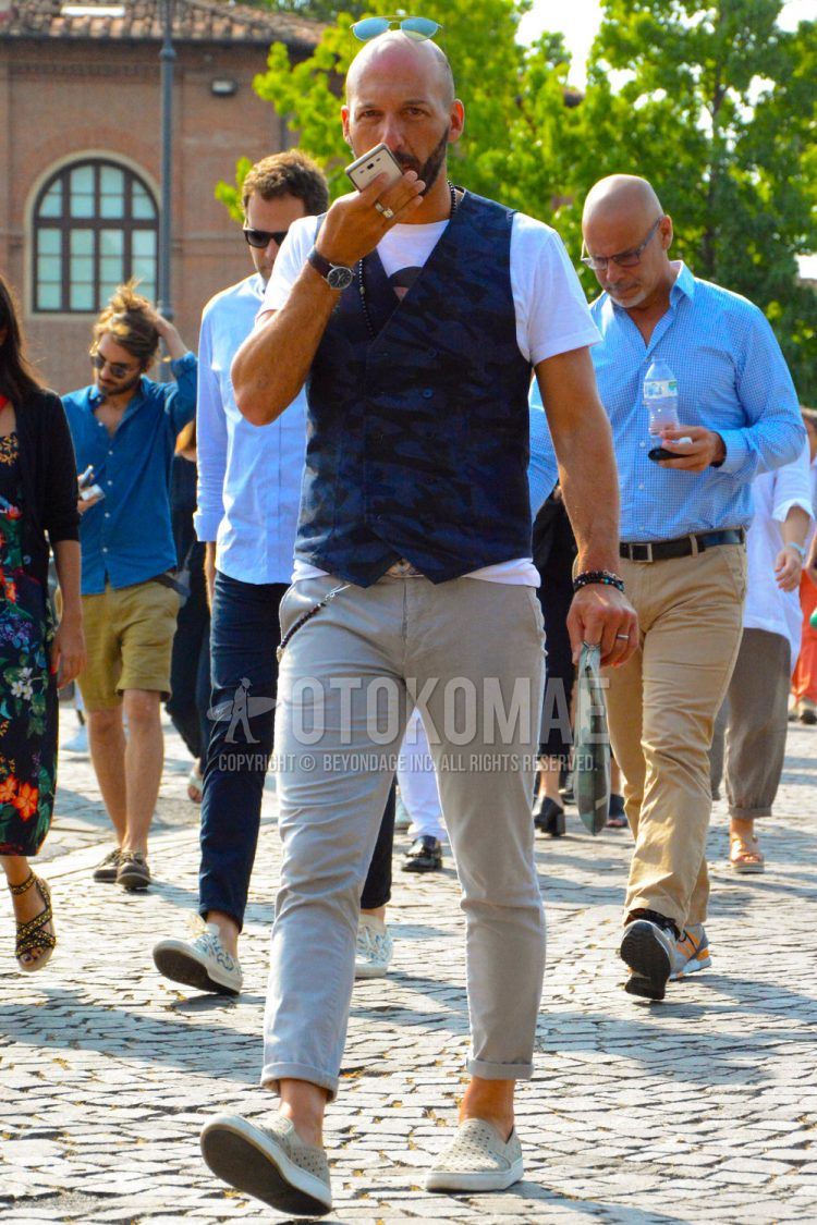 A summer men's coordinate outfit with a navy camouflage gilet, plain white t-shirt, plain beige chinos, and beige slip-on sneakers.