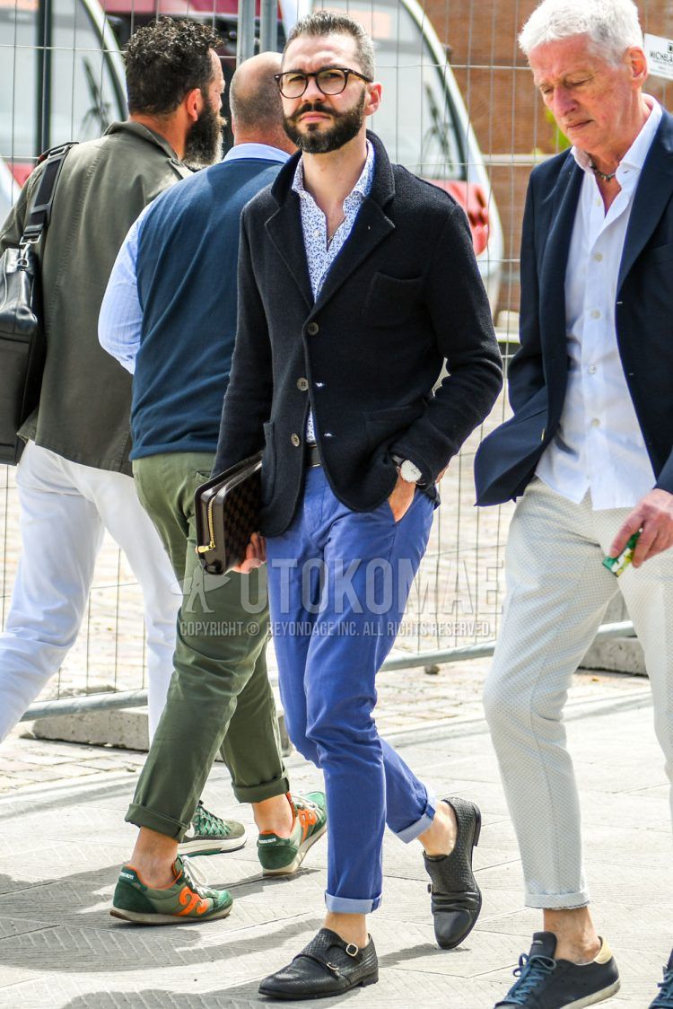 A summer/spring/fall men's outfit with plain black sunglasses, plain black tailored jacket, white/blue or other shirt, plain blue/light blue chinos, black monk shoe leather shoes, and plain black clutch/second bag/drawstring bag.