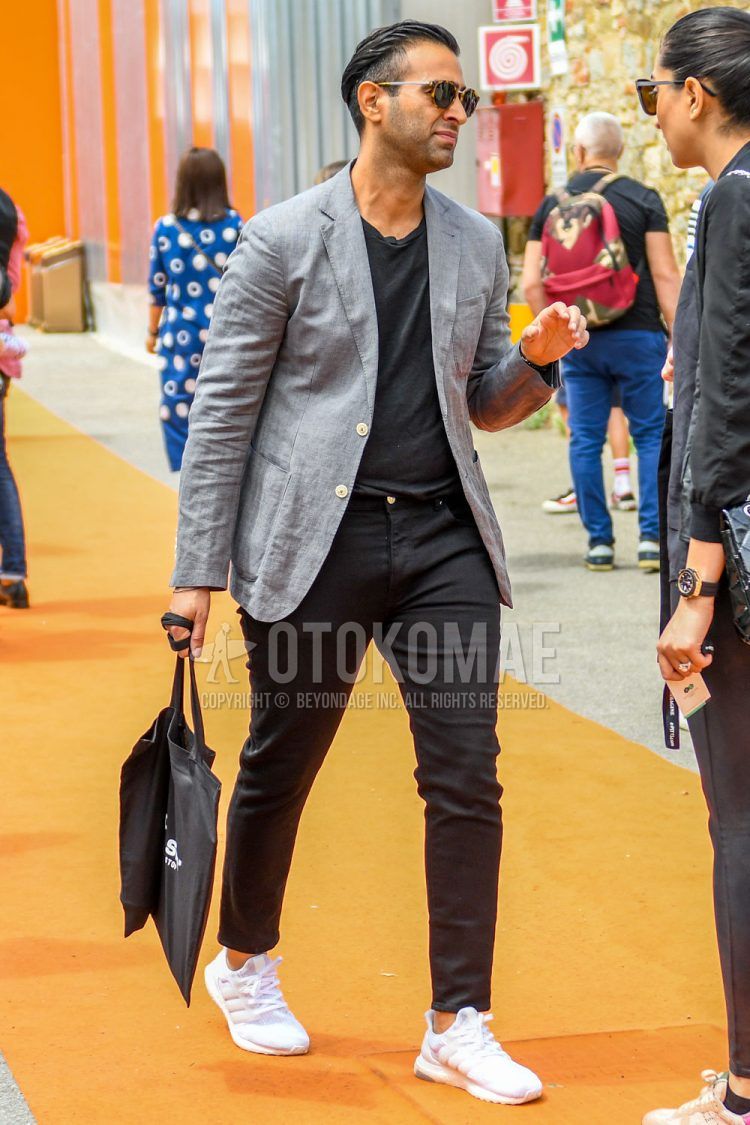 A men's spring/summer/fall outfit of plain sunglasses, plain gray tailored jacket, plain black t-shirt, plain black ankle pants, plain black denim/jeans, and white low-cut Adidas sneakers.