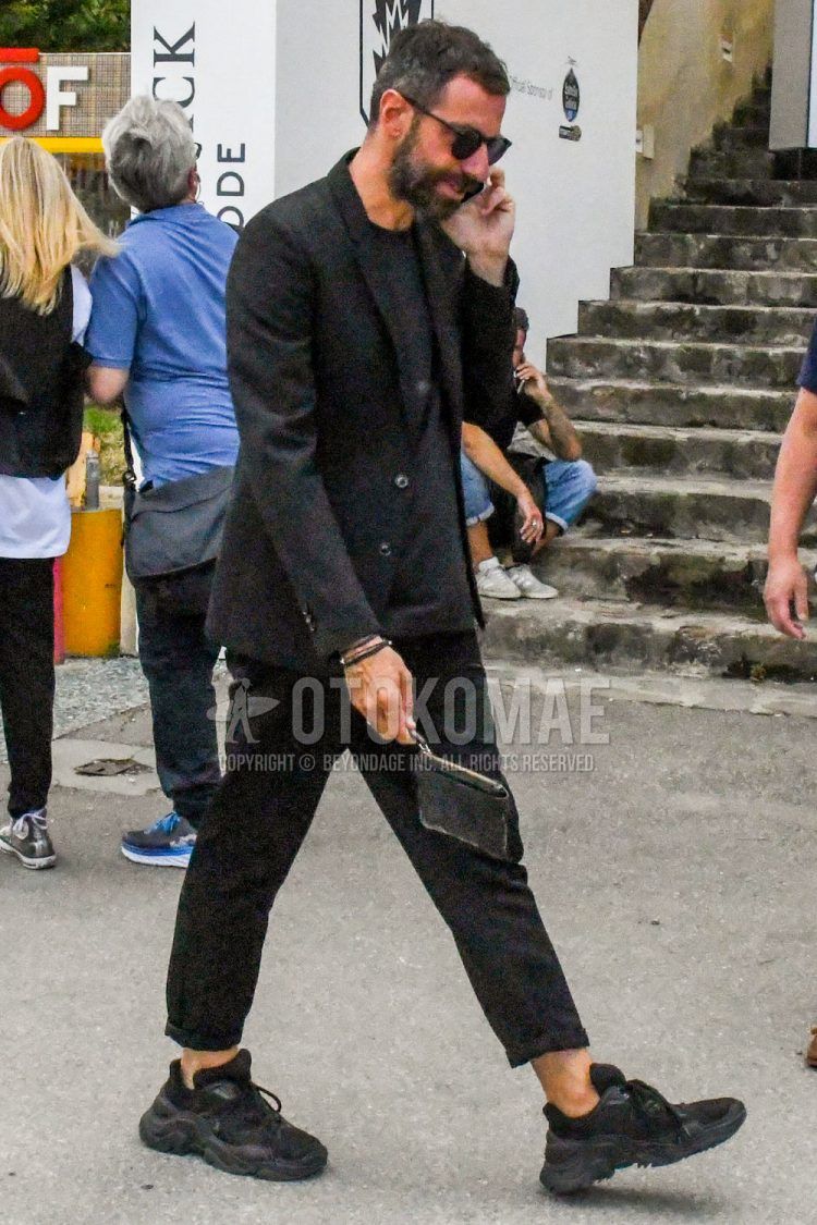 A men's spring/fall/summer outfit for men with solid color sunglasses, solid color black tailored jacket, solid color black t-shirt, solid color black cropped pants, black low-cut sneakers, and solid color black clutch/second bag/drawstring bag.