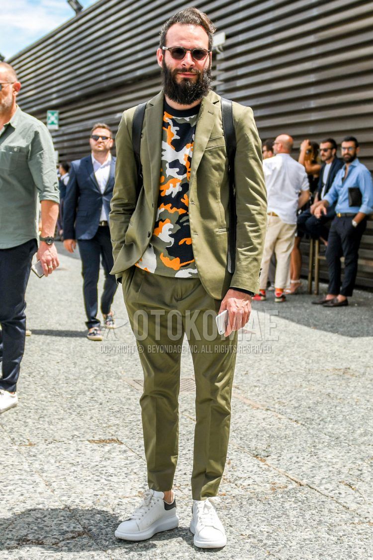 A summer/fall/spring men's coordinate outfit with plain sunglasses, a multi-colored camouflage t-shirt, Alexander McQueen white low-cut sneakers, and a plain olive green suit.
