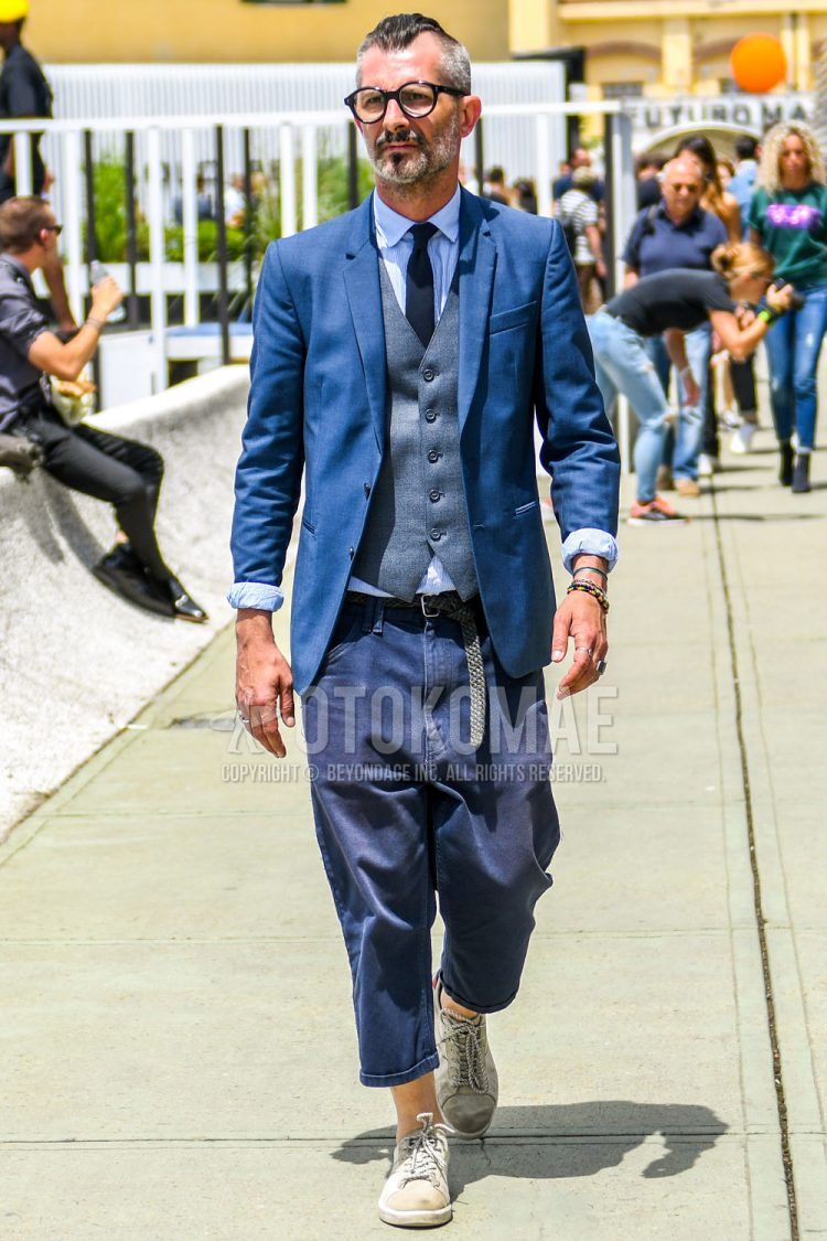 A summer-spring-fall men's outfit with plain glasses, plain blue tailored jacket, plain gray gilet, white striped shirt, plain black mesh belt, plain navy sarouel pants, beige low-cut sneakers, and plain navy tie.