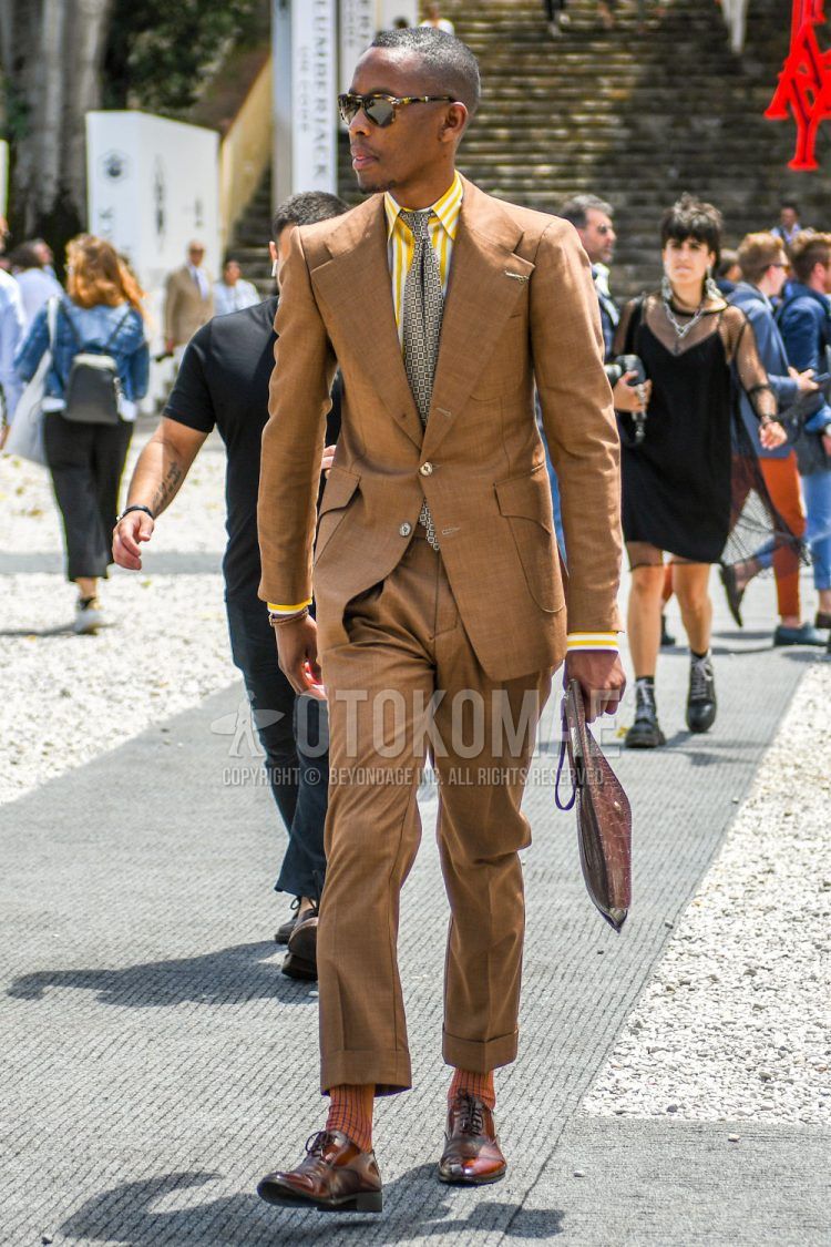 Summer/spring/fall men's coordinate outfit with tortoiseshell sunglasses, yellow/white striped shirt, orange striped socks, brown straight tip leather shoes, brown solid color clutch bag/second bag/drawstring, beige solid color suit, brown small print tie.
