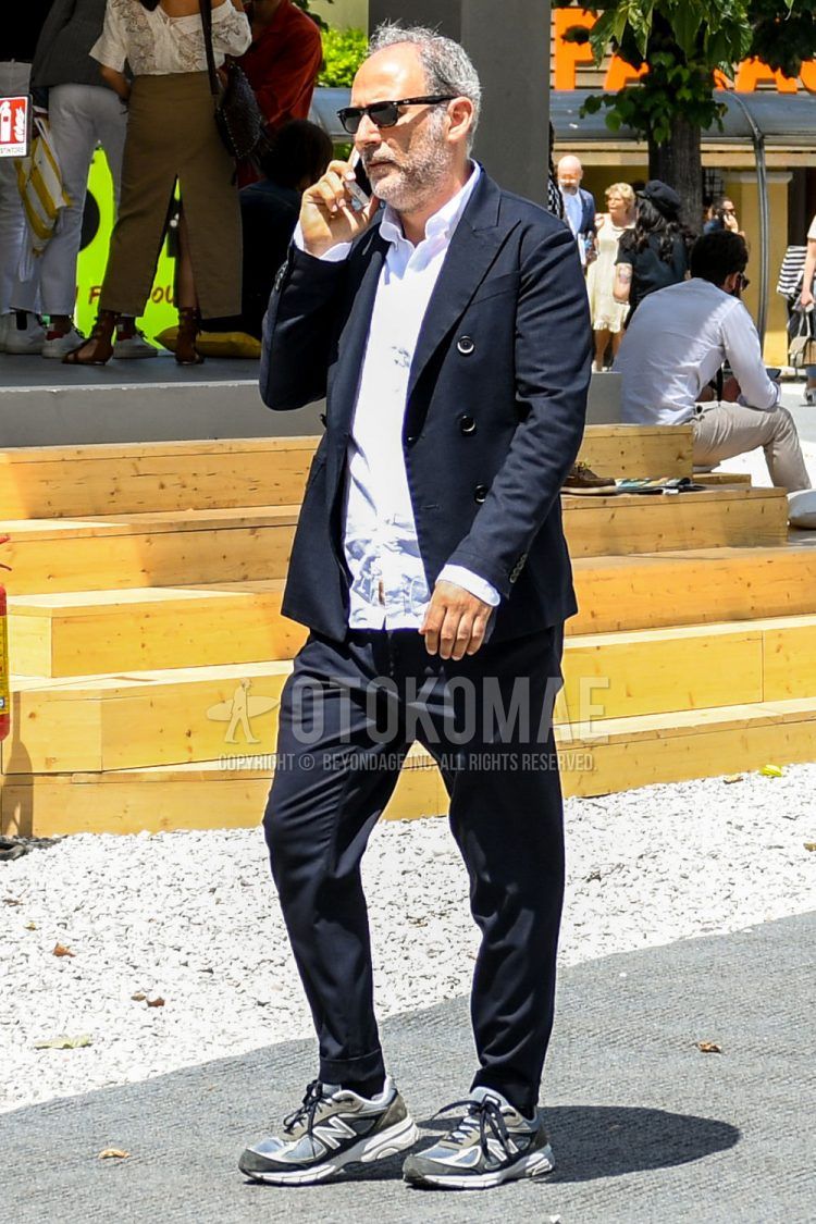 A men's spring/summer/fall outfit for men with plain sunglasses, a plain white shirt, New Balance gray/navy low-cut sneakers, and a plain black suit.