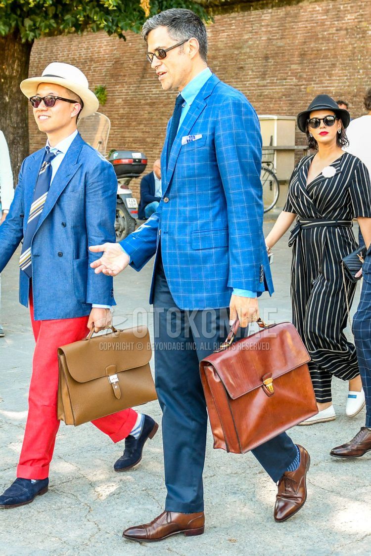 Spring/Summer/Fall men's outfit with navy check tailored jacket, light blue solid shirt, navy solid slacks, blue striped socks, brown straight tip leather shoes, brown solid briefcase/handbag.