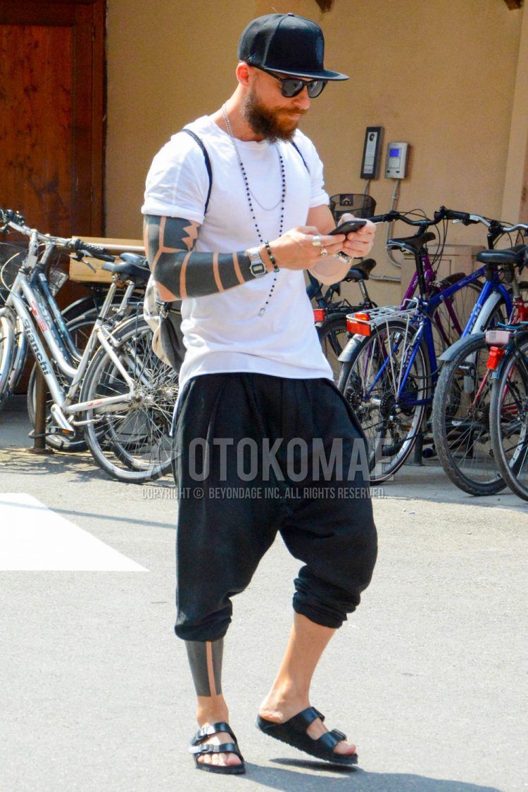 A men's spring/summer coordinate outfit with a solid color baseball cap, solid color sunglasses, solid color white t-shirt, solid color black sarouel pants, and black leather sandals.