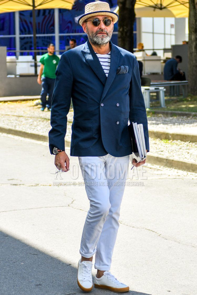 A men's spring/summer/fall outfit for men with a plain hat, plain sunglasses, plain navy tailored jacket, white striped t-shirt, plain white denim/jeans, and white low-cut sneakers.