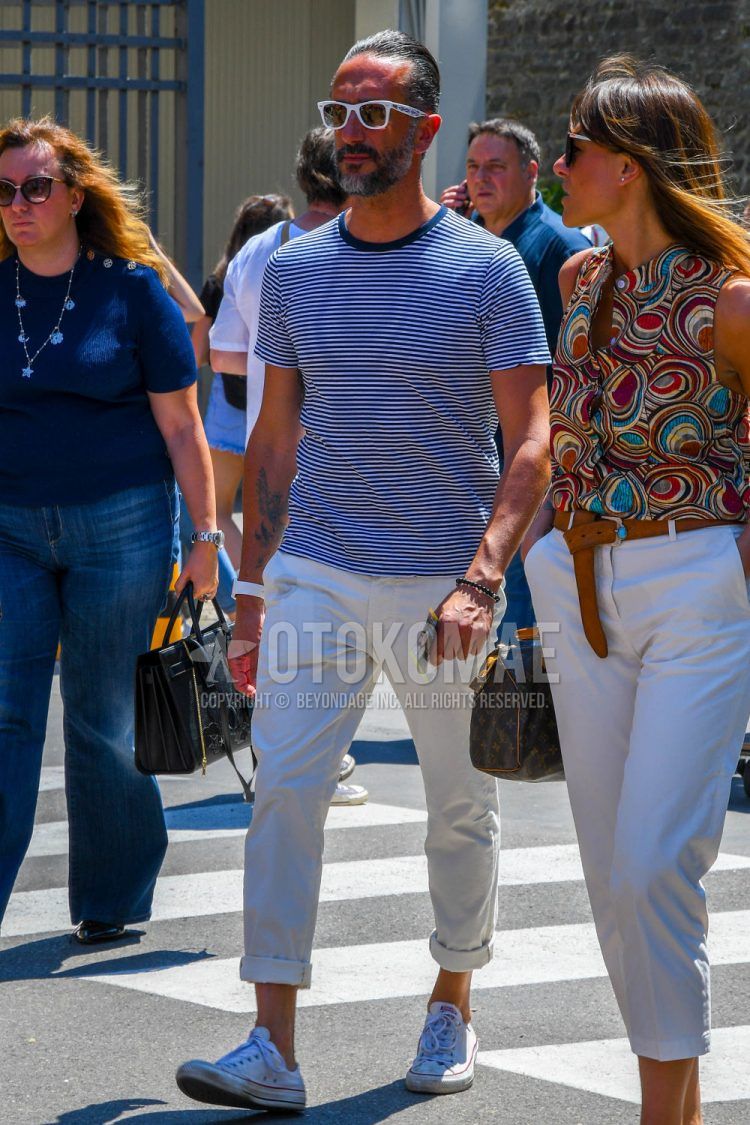 A summer men's coordinate outfit with plain glasses, a white/navy striped t-shirt, plain beige cotton pants, and white low-cut Converse Chuck Taylor sneakers.