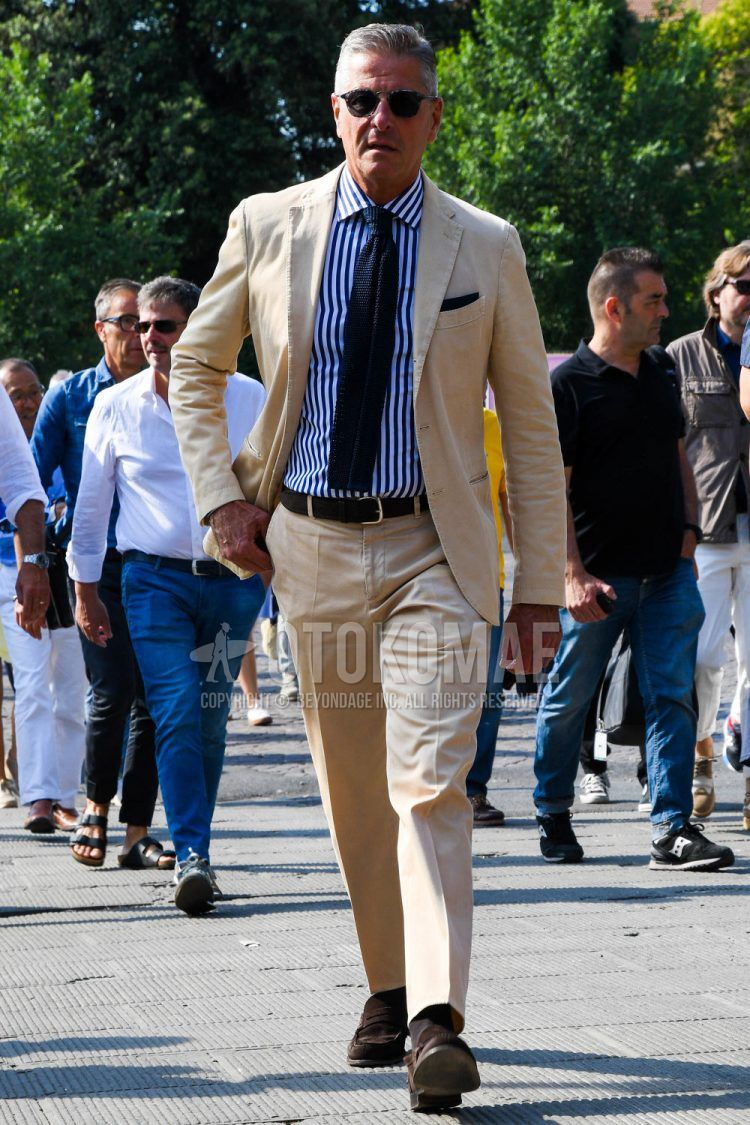 Spring, summer and fall men's coordinate outfit with plain sunglasses, white striped shirt, plain leather belt, brown coin loafers leather shoes, brown suede shoes leather shoes, plain beige suit and plain navy knit tie.