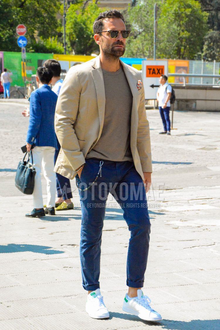 Spring, summer and fall men's coordinate outfit with tortoiseshell sunglasses, plain beige tailored jacket, plain beige t-shirt, plain blue chinos and Adidas Stan Smith white low-cut sneakers.