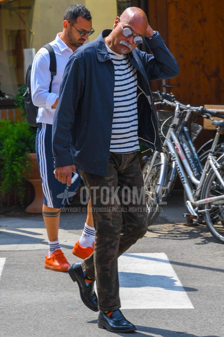 Spring, summer and fall men's coordinate outfit with solid color sunglasses, navy dot coach jacket, white striped t-shirt, olive green camouflage chinos and black coin loafer leather shoes.