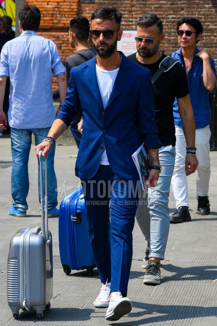 Spring, summer and fall men's coordinate outfit with plain black sunglasses, plain white T-shirt, white low-cut Converse sneakers, plain silver suitcase and blue/navy check suit.