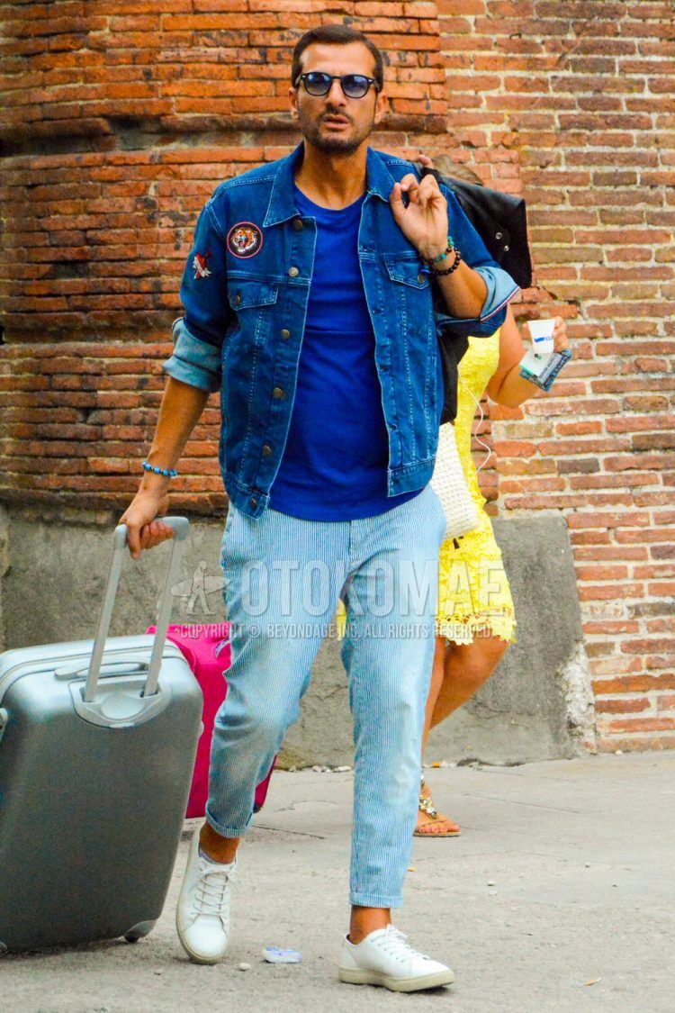 Spring, summer and fall men's coordinate outfit with solid color sunglasses, solid color blue denim jacket, solid color blue t-shirt, blue striped cotton pants, white low-cut sneakers and solid color gray suitcase.
