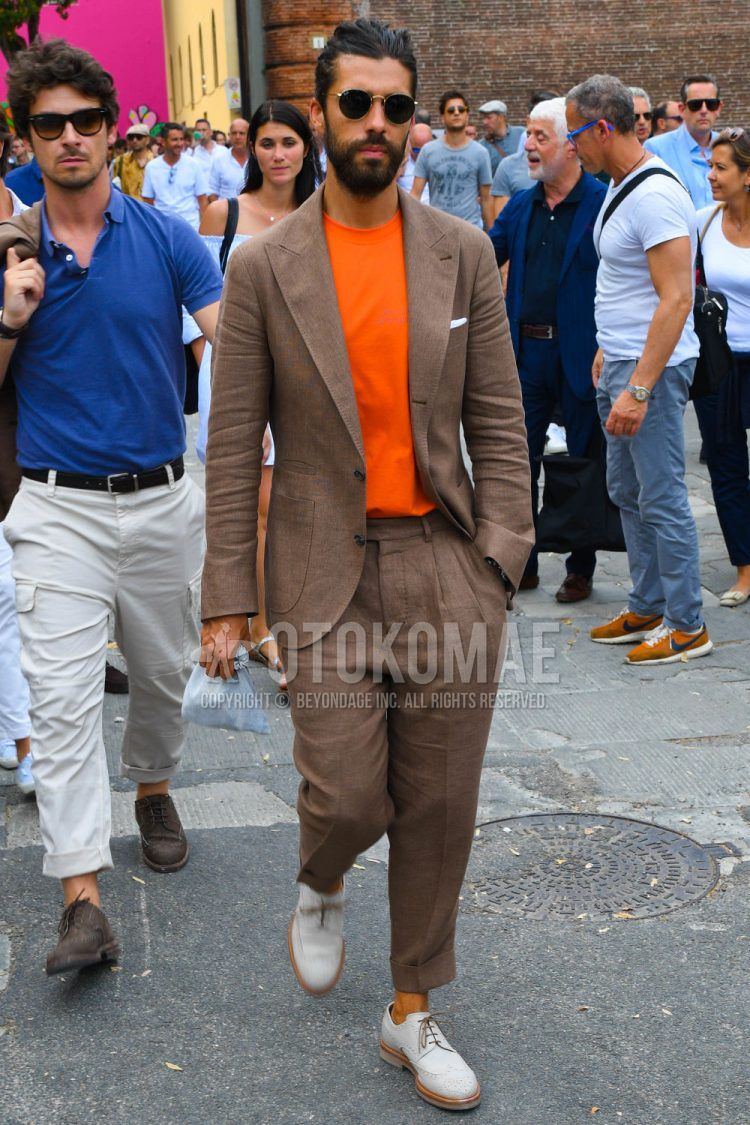 A men's spring/summer/fall outfit for men with plain sunglasses, plain orange t-shirt, leather shoes with white brogue shoes, and plain brown suit.