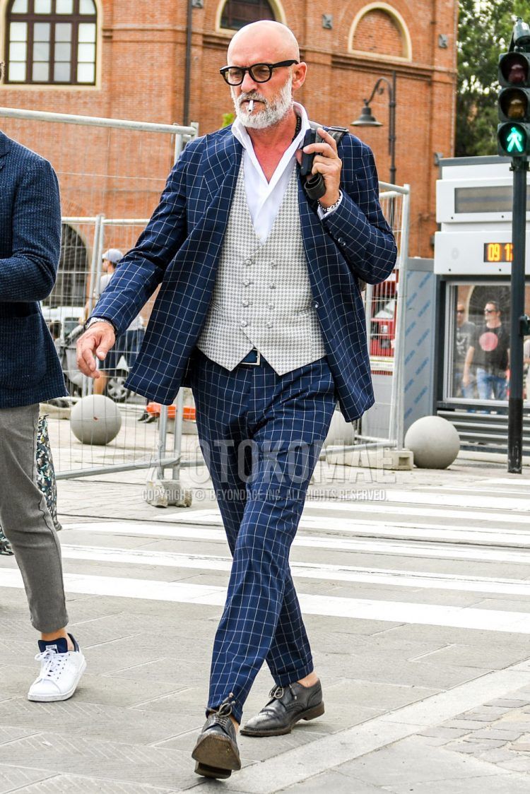 A summer/fall/spring men's coordinate outfit with plain glasses, gray checked gilet, plain white shirt, plain blue leather belt, black brogue shoes leather shoes, and navy check suit.