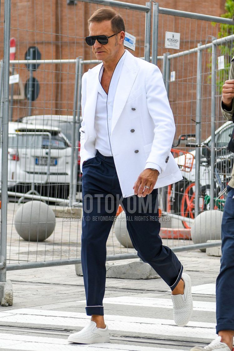 A men's spring, fall, and summer outfit with plain sunglasses, a plain white tailored jacket, a plain white shirt, plain navy slacks, and white slip-on sneakers.