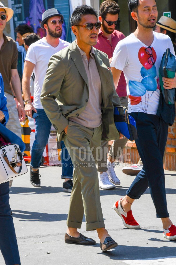 A men's spring/fall/summer coordinate outfit with tortoiseshell sunglasses, plain gray polo shirt, brown other loafer leather shoes, plain clutch bag/second bag/drawstring, and plain olive green suit.