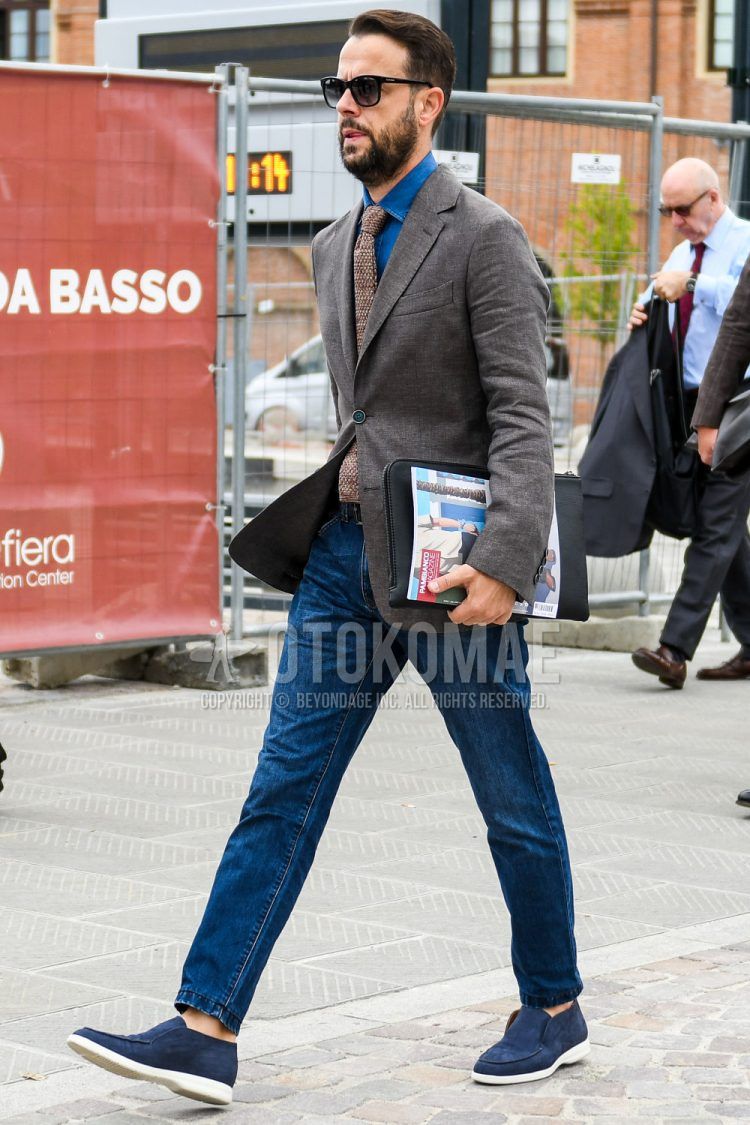 Solid color sunglasses, solid color brown tailored jacket, solid color blue denim/chambray shirt, solid color blue denim/jeans, blue low cut sneakers, solid color black clutch bag/second bag/drawstring, solid color brown knit tie, spring/summer/fall men's coordinate Outfit.