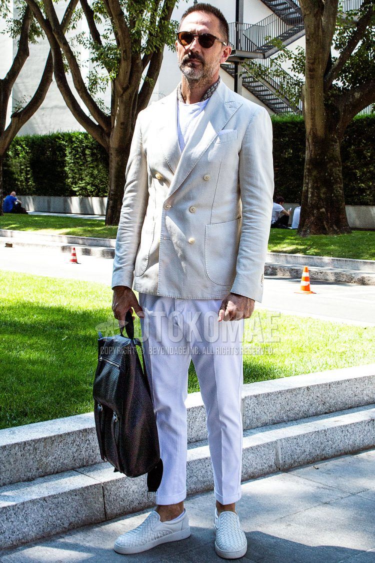 Spring and fall men's outfit with plain sunglasses, graphic bandana/neckerchief, plain white tailored jacket, plain white t-shirt, plain white slacks, white slip-on sneakers, and plain brown backpack from Bottega Veneta.