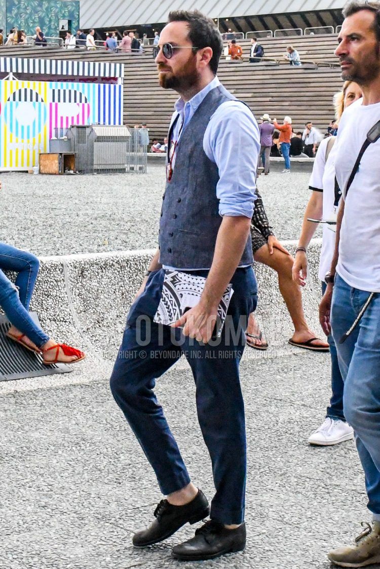 A men's spring/summer outfit with solid color sunglasses, solid color blue shirt, solid color gray gilet, solid color navy slacks, and black plain toe leather shoes.